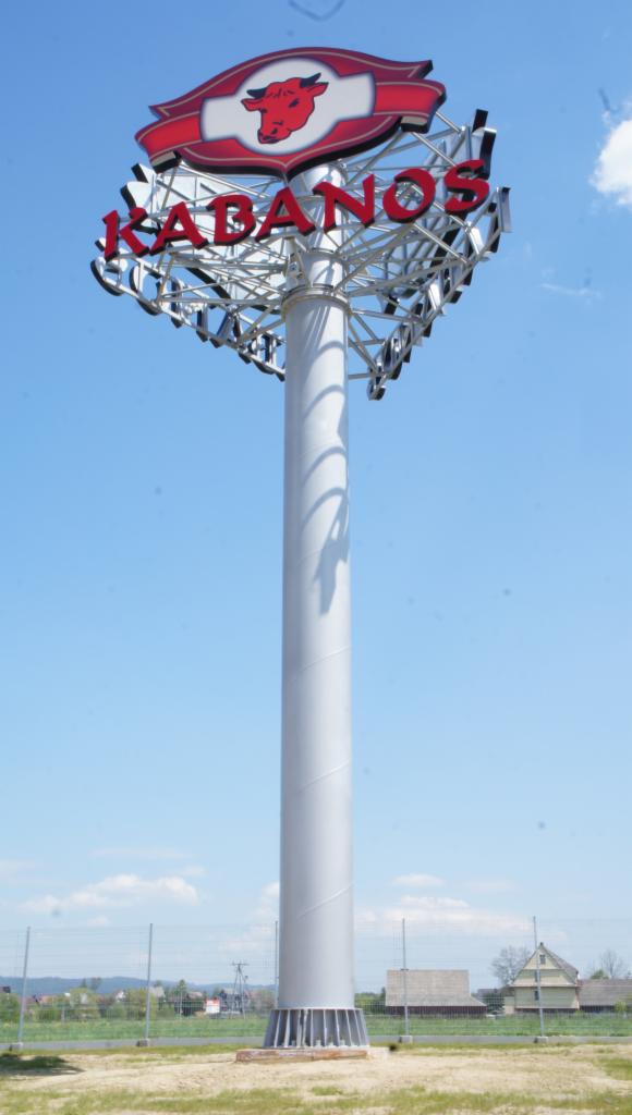Wysoki pylon reklamowy marki Kabanos z czerwonym logo byka, umieszczony na metalowej konstrukcji na tle błękitnego nieba i zabudowań w oddali.