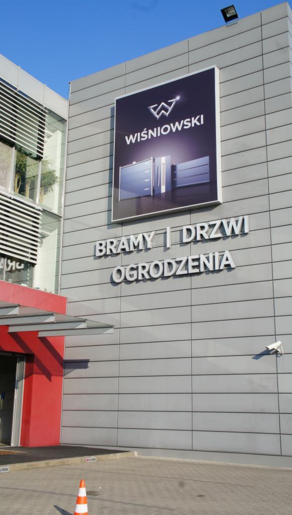 Elewacja budynku z dużym banerem firmy Wiśniowski, oferującej bramy, drzwi i ogrodzenia. Widoczny fragment wejścia do budynku i pomarańczowy pachołek.