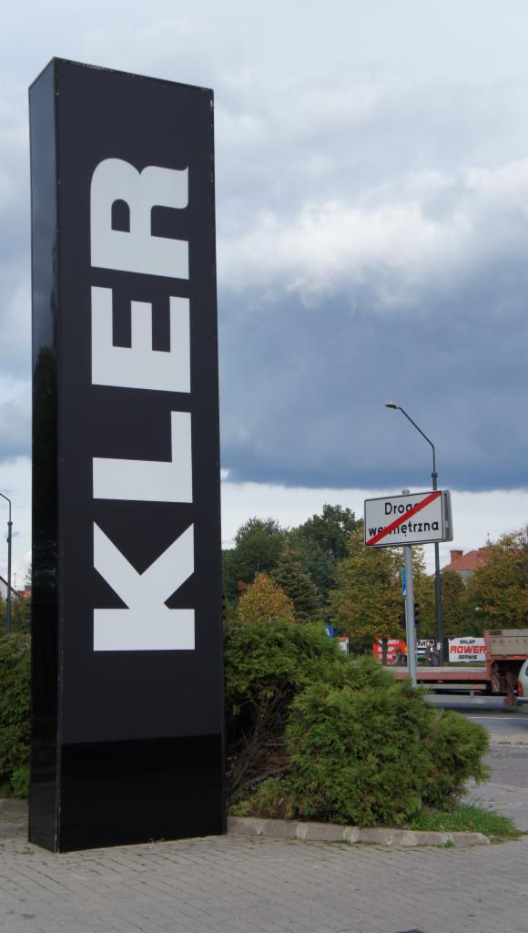 Wysoki, czarny pylon reklamowy z białym napisem 'KLER' na tle pochmurnego nieba i zieleni miejskiej. W tle znak drogowy 'Droga wewnętrzna'.