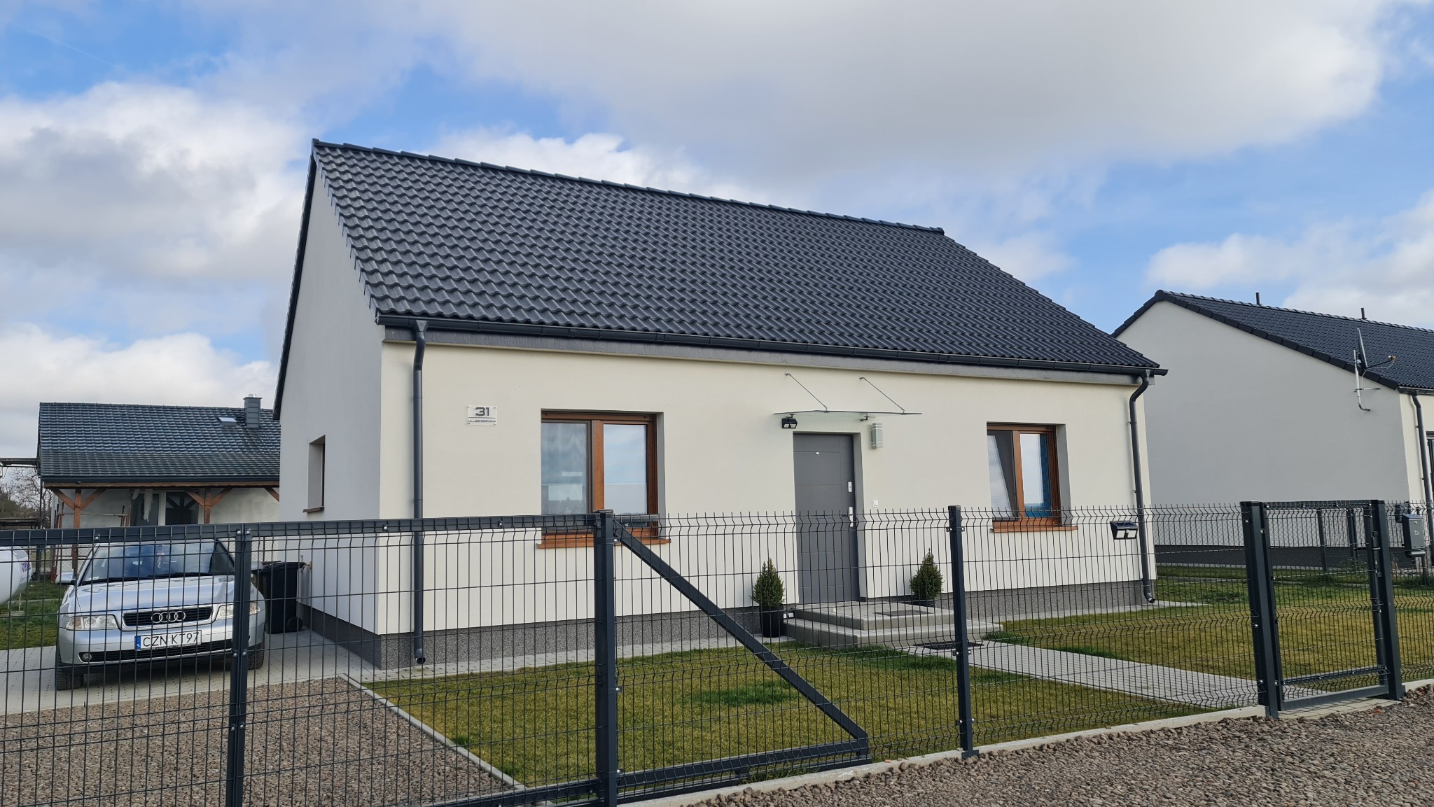 Nowoczesny dom murowany z ciemną dachówką, widok zza ogrodzenia, z numerem 31 na elewacji.