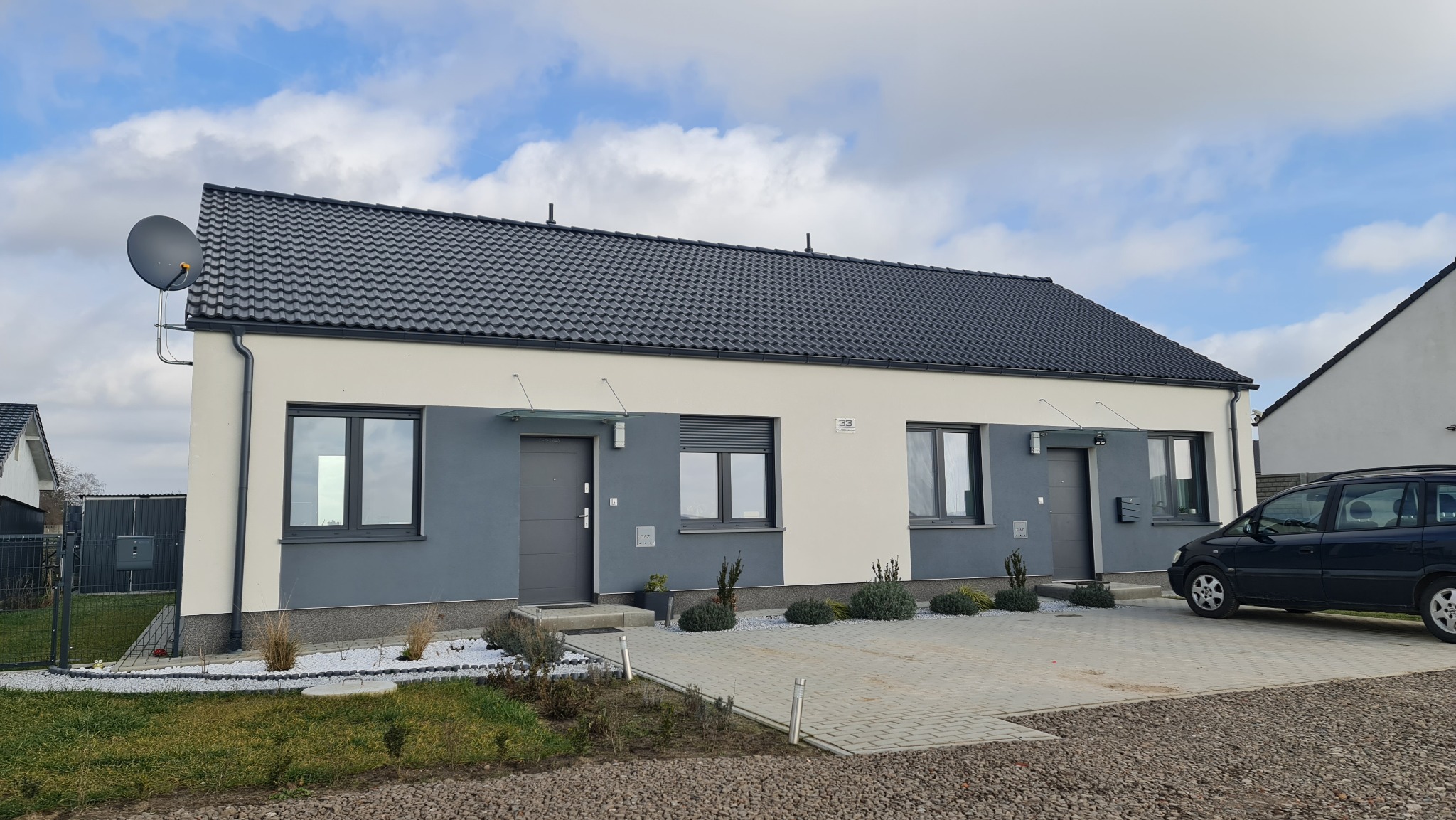 Dwa identyczne domy jednorodzinne w zabudowie bliźniaczej z szarymi dachami i elewacją w odcieniach szarości i bieli, z satelitą na jednym z budynków i zaparkowanym samochodem osobowym przed drugim.