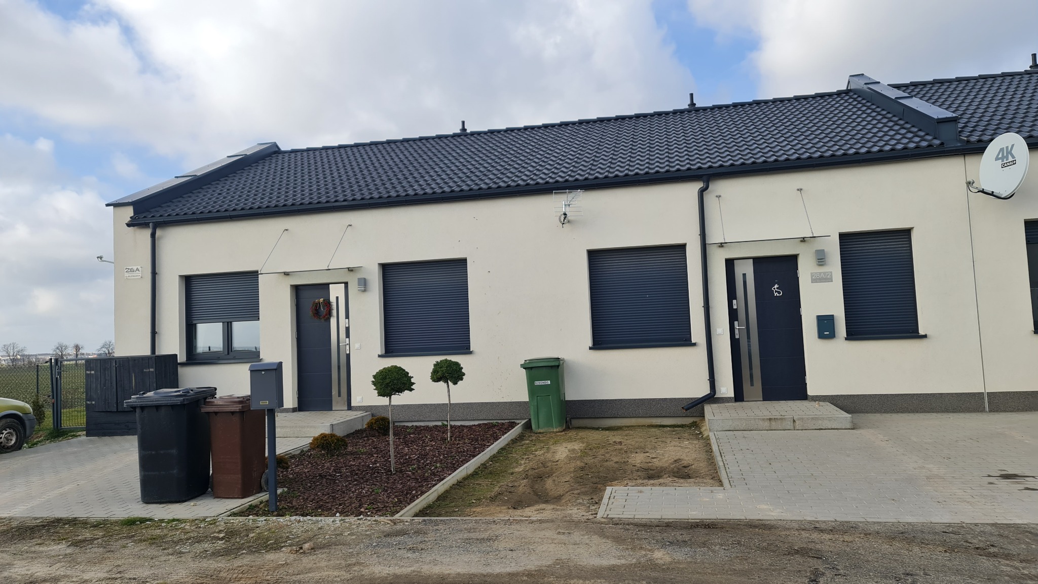 Bliźniak z jasną elewacją, ciemną dachówką i roletami zewnętrznymi, widoczne kosze na śmieci i małe drzewka ozdobne.