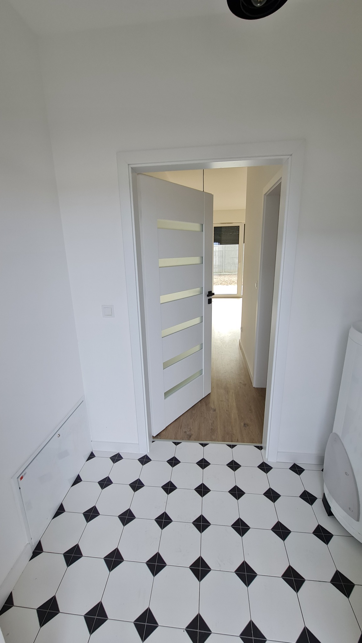 Wnętrze domu w stanie deweloperskim: białe ściany, podłoga w biało-czarną szachownicę, częściowo otwarte białe drzwi z poziomymi przeszkleniami prowadzące do pomieszczenia z drewnianą podłogą.