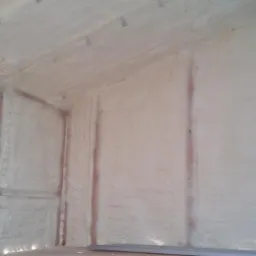 Wnętrze pomieszczenia z drewnianą konstrukcją szkieletową, pokryte warstwą izolacyjnej piany o nierównej powierzchni, widoczne fragmenty okna.