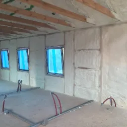 Wnętrze budynku w trakcie prac izolacyjnych, ściany i sufit pokryte białą pianą, okna zabezpieczone niebieską folią, widoczne instalacje podłogowe.