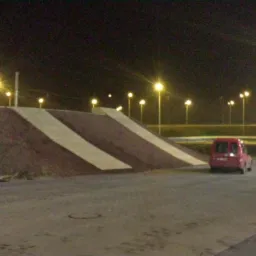 Nocny widok na ziemny nasyp z betonowymi pasami, oświetlony latarniami, z czerwonym samochodem w tle i fragmentem czarnego auta po prawej.