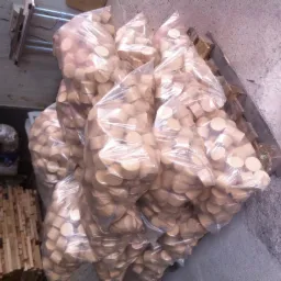 Stos worków foliowych wypełnionych okrągłymi kawałkami drewna na palecie, obok rury izolacyjnej i ułożonych listewek drewnianych.