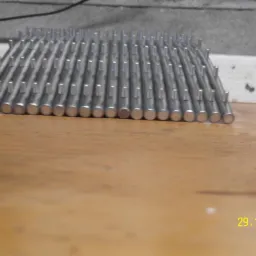 Metalowa konstrukcja z rzędami równo rozmieszczonych, okrągłych prętów na drewnianej powierzchni, widziana z niskiej perspektywy.