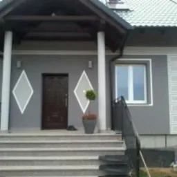 Szary dom jednorodzinny z ciemnymi elementami wykończeniowymi, widoczne schody wejściowe z szarej kostki, elewacja z dekoracyjnymi rombami i pionowymi pasami, małe drzewko w donicy przed wejściem.