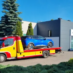 Żółto-czerwona laweta z niebieskim Peugeotem na lawecie, na tle salonu Skody. Samochód zabezpieczony pasami transportowymi. Słoneczny dzień.