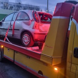 TS24 Pomoc Drogowa Jastrowie - Uszkodzony, srebrny Opel Corsa na żółtej lawecie TEVOR, z widocznymi uszkodzeniami karoserii i wybitymi szybami, po zmroku, na tle zielonego ekranu akustycznego.
