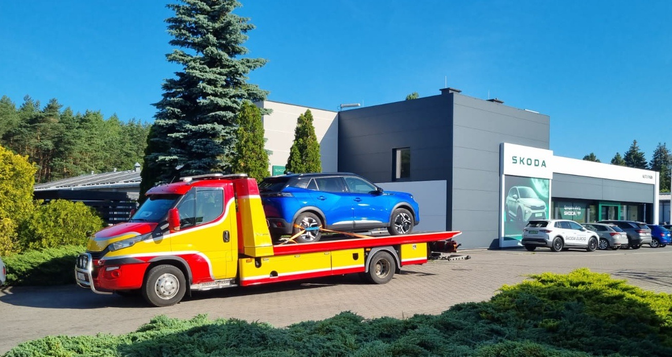 Żółto-czerwona laweta z niebieskim Peugeotem na lawecie, na tle salonu Skody. Samochód zabezpieczony pasami transportowymi. Słoneczny dzień.