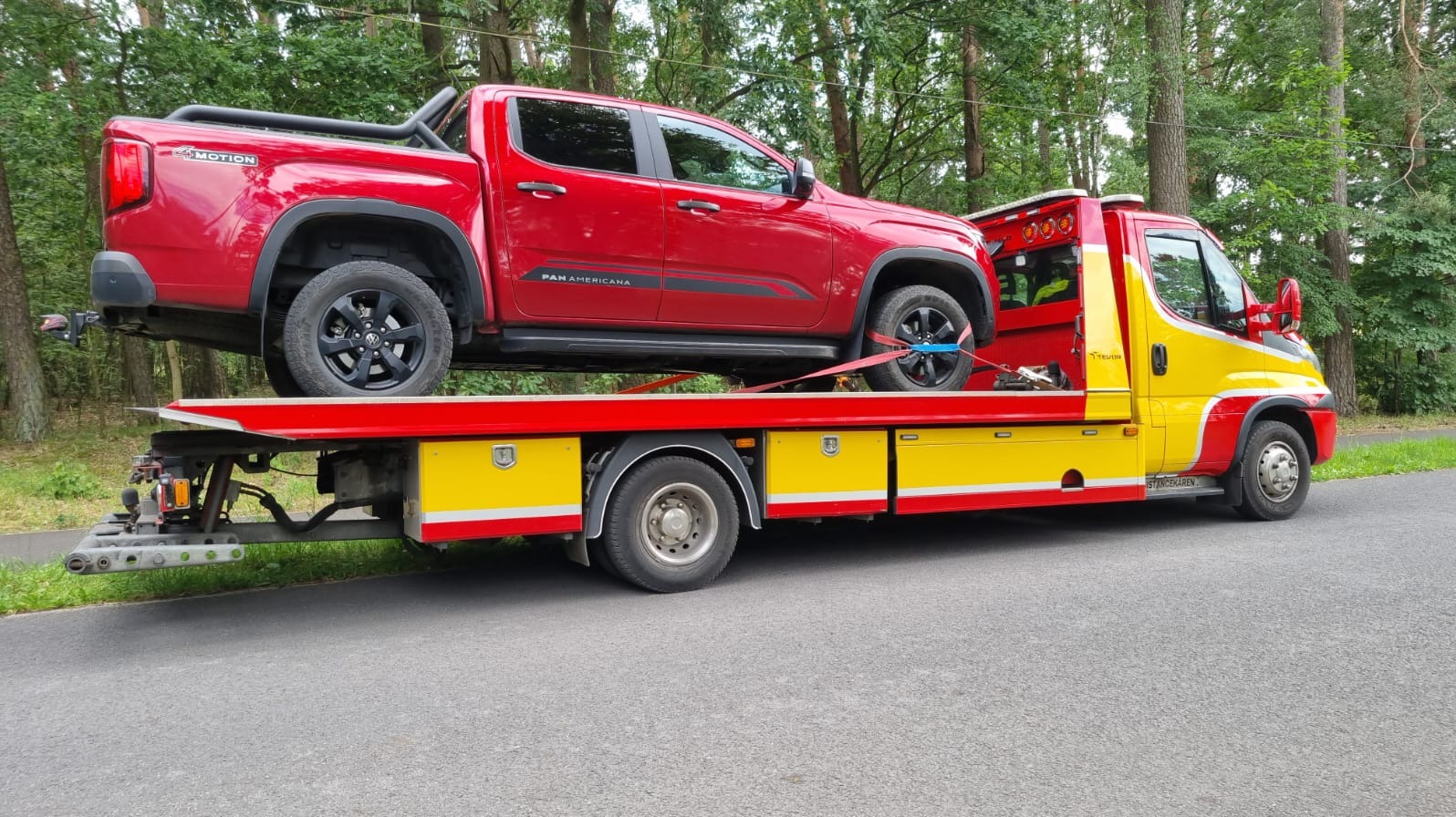 Czerwony pickup 'Panamericana' na żółto-czerwonej lawecie, zabezpieczony pasami transportowymi, na tle zieleni. Usługa transportowa.