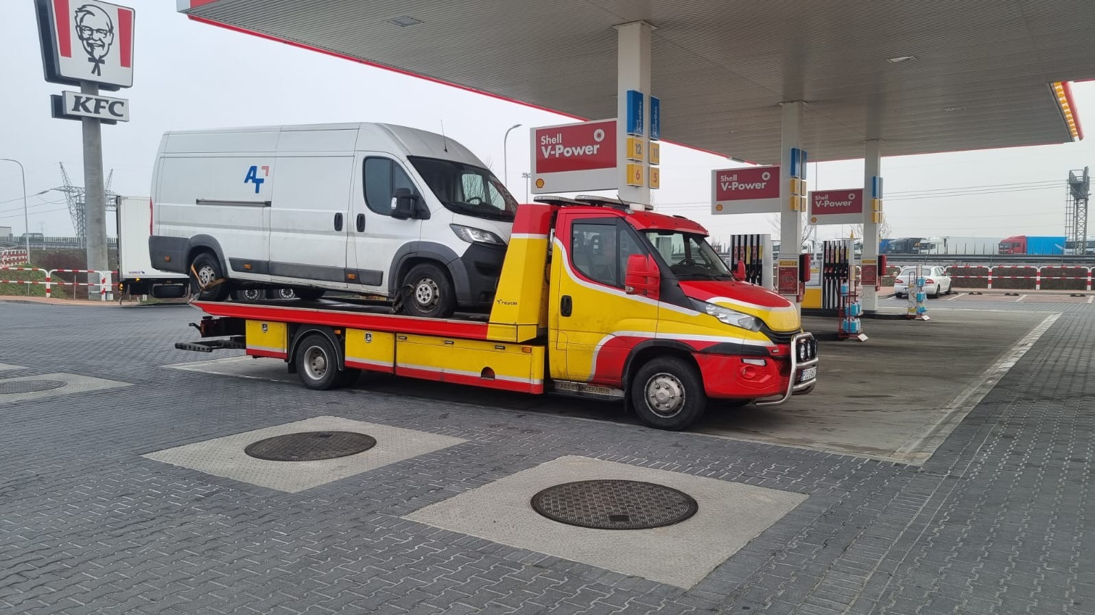 Żółto-czerwona laweta z białym busem na platformie, stojąca na stacji benzynowej Shell z reklamą KFC w tle. Transport awaryjny pojazdu.