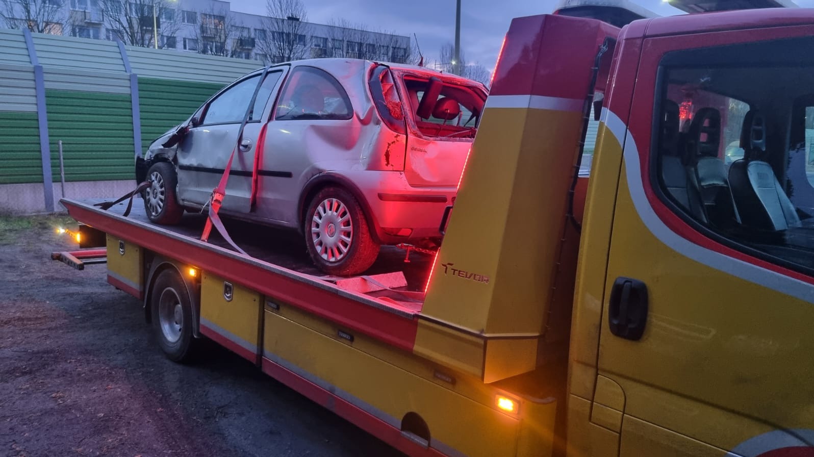 Uszkodzony, srebrny Opel Corsa na żółtej lawecie TEVOR, z widocznymi uszkodzeniami karoserii i wybitymi szybami, po zmroku, na tle zielonego ekranu akustycznego.