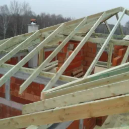 Konstrukcja drewniana więźby dachowej na murowanym budynku w trakcie budowy, widoczne krokwie, murłaty i komin w tle.