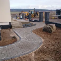 Kaskadowa ścieżka z kostki brukowej prowadząca do domu, z elementami dekoracyjnymi i ciężkim sprzętem budowlanym w tle, podkreślająca kompleksowość prac brukarskich.