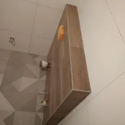 Wykończona łazienka w Rawiczu: zabudowa z drewnopodobnych płytek nad toaletą, z detalami w postaci małej półki z figurką i uchwytu na papier toaletowy, na tle ściany z geometrycznym wzorem płytek.