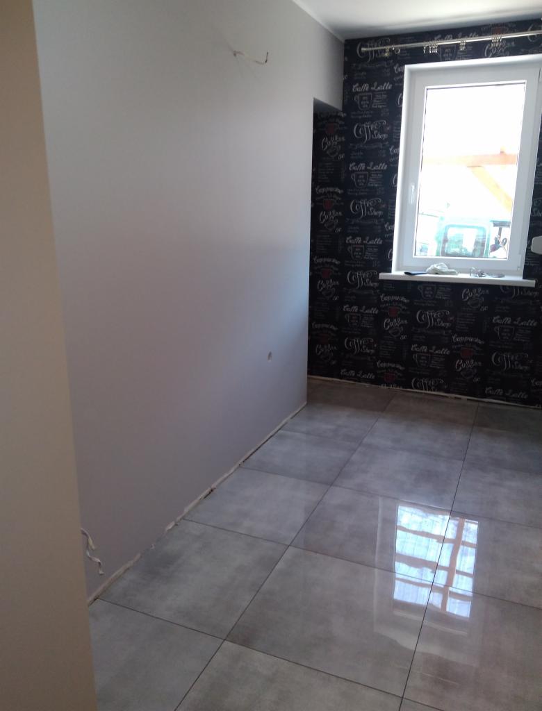 Pokój po remoncie z szarą ścianą, tapetą z motywem kawowym i szarą podłogą z płytek. Widoczne ślady po usuwaniu starych listew przypodłogowych.