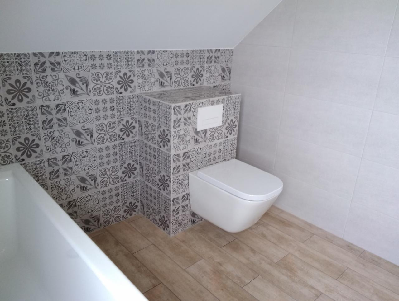 Wykończona łazienka na poddaszu: wzorzyste płytki ceramiczne na ścianie za toaletą i częściowo nad wanną, kontrastujące z gładkimi, białymi płytami po prawej stronie i panelami podłogowymi...