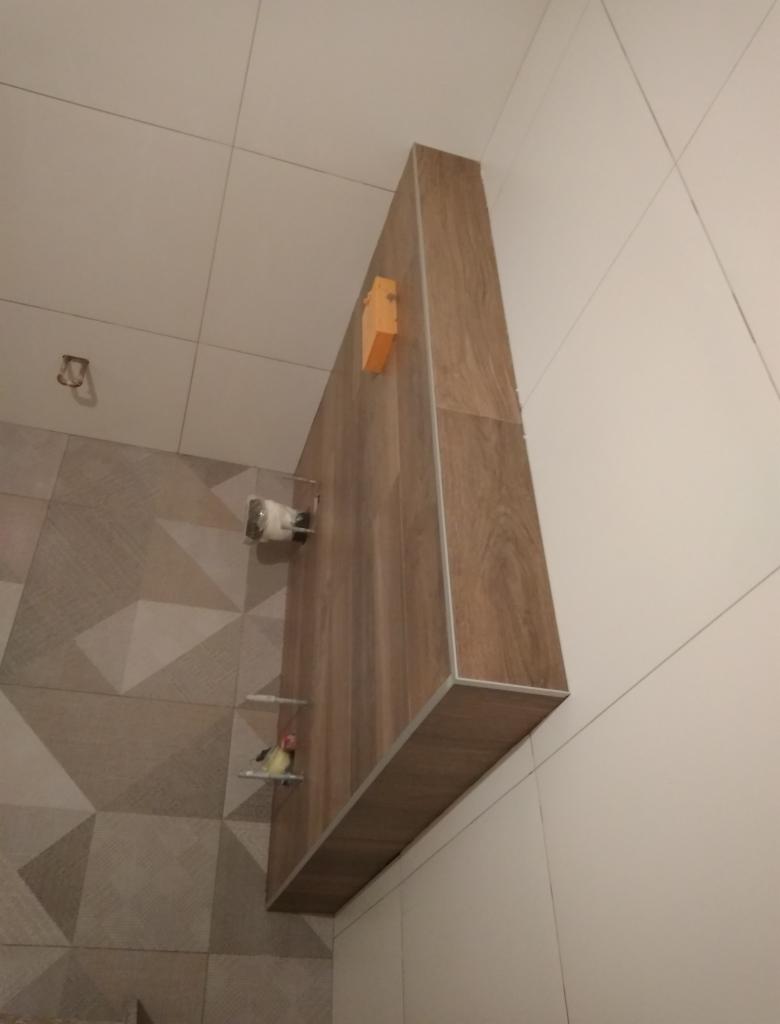 Wykończona łazienka w Rawiczu: zabudowa z drewnopodobnych płytek nad toaletą, z detalami w postaci małej półki z figurką i uchwytu na papier toaletowy, na tle ściany z geometrycznym wzorem płytek.