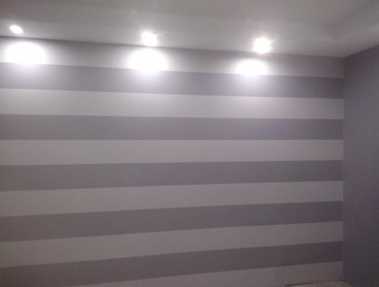 Pomieszczenie z pomalowaną ścianą w poziome, szare pasy; trzy lampy sufitowe oświetlają górną część ściany.