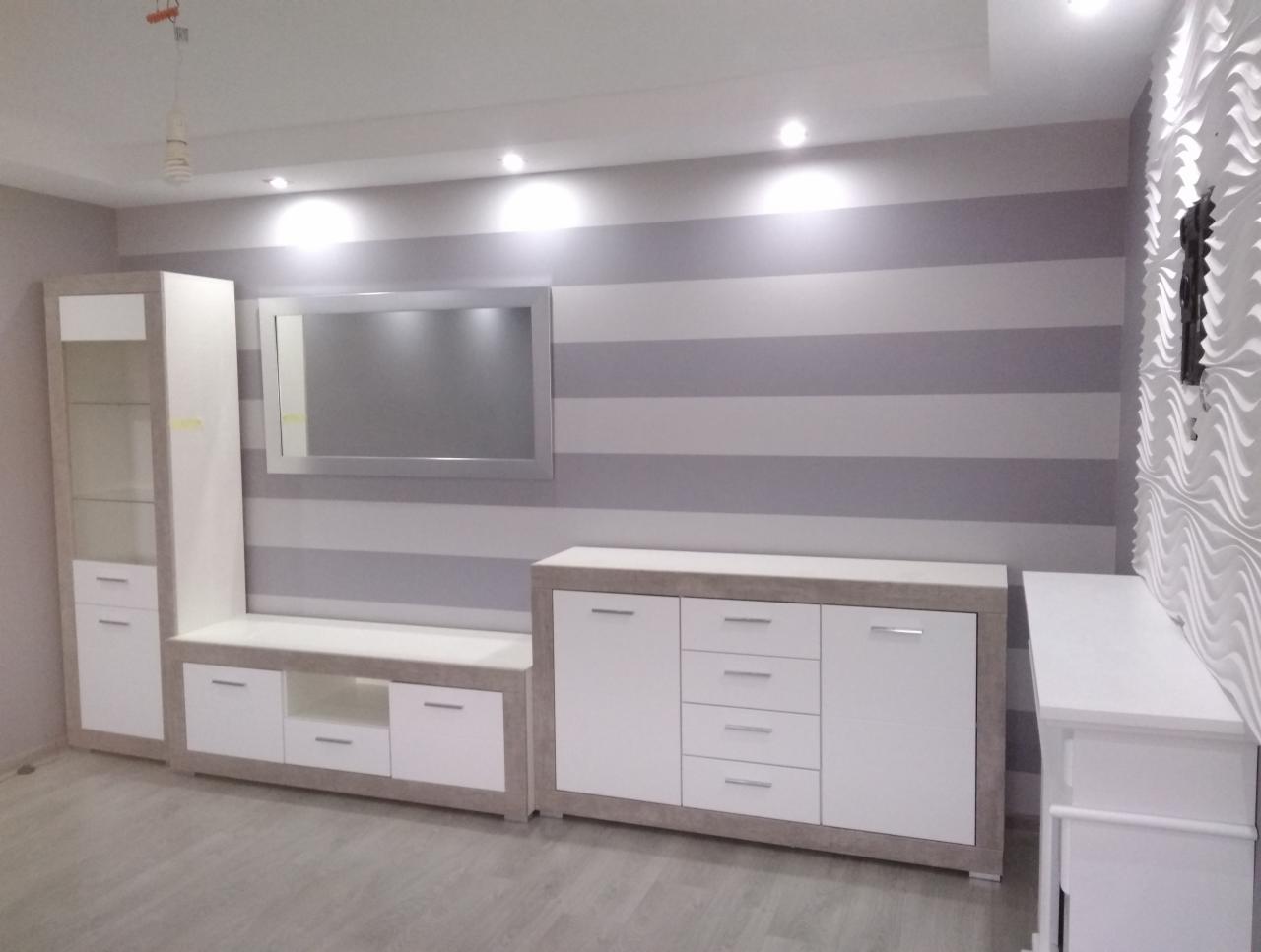 Nowoczesny salon z białymi meblami z akcentami w kolorze jasnego drewna, szarą ścianą w poziome pasy i dekoracyjną białą płaskorzeźbą na ścianie po prawej stronie.