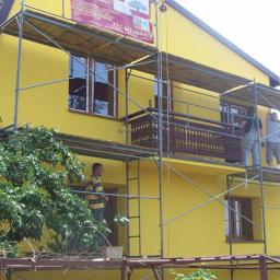 Reno-Bud - Żółty dom w trakcie malowania elewacji. Widoczne rusztowania i ekipa malarzy pracująca na wysokości. Na rusztowaniu baner reklamowy firmy.