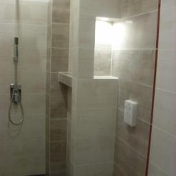 Reno-Bud - Wykończona łazienka z beżowymi płytkami, wnęką z półką i deszczownicą. Widoczne elementy instalacji wodnej i gniazdko elektryczne. Minimalistyczny design.