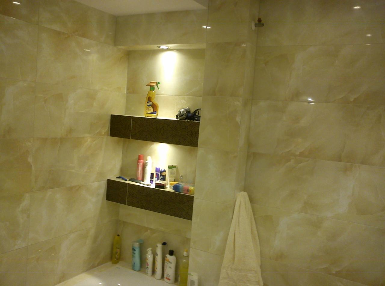 Wykończona łazienka z beżowymi płytkami imitującymi marmur. Wnęka z półkami podświetlona punktowym światłem. Widoczne kosmetyki i akcesoria kąpielowe.