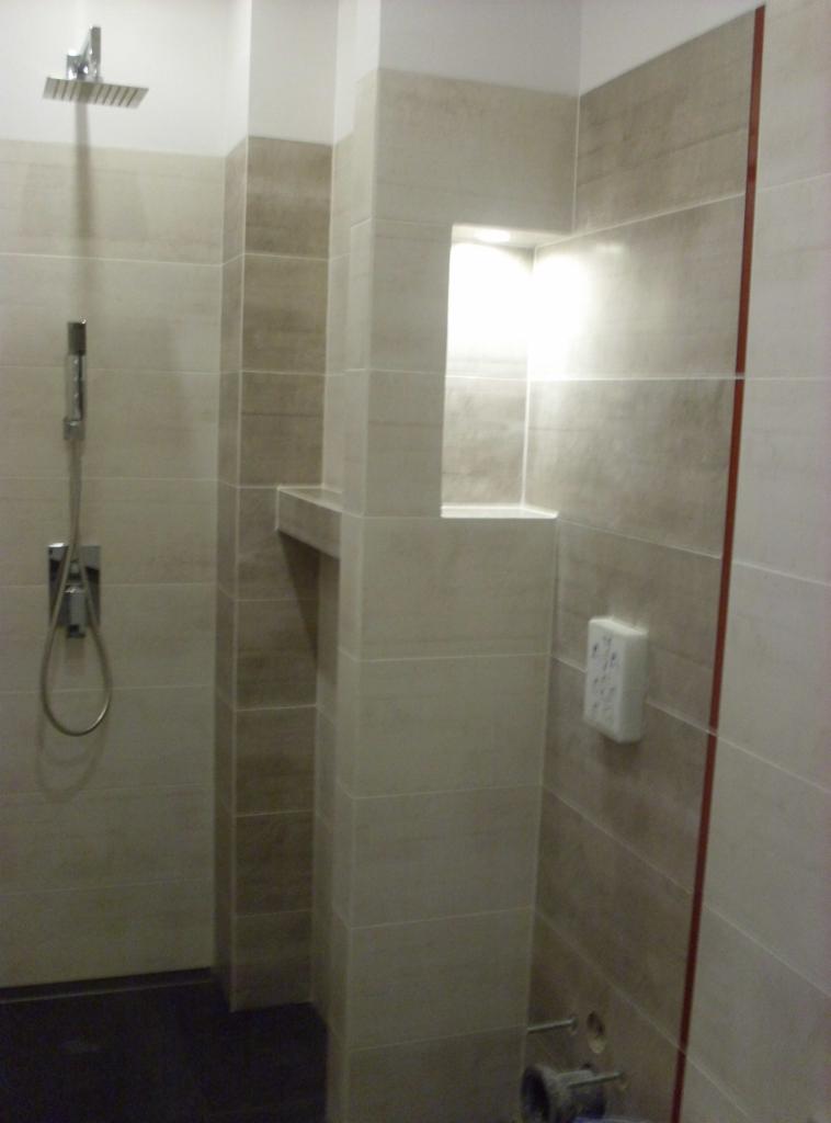 Wykończona łazienka z beżowymi płytkami, wnęką z półką i deszczownicą. Widoczne elementy instalacji wodnej i gniazdko elektryczne. Minimalistyczny design.