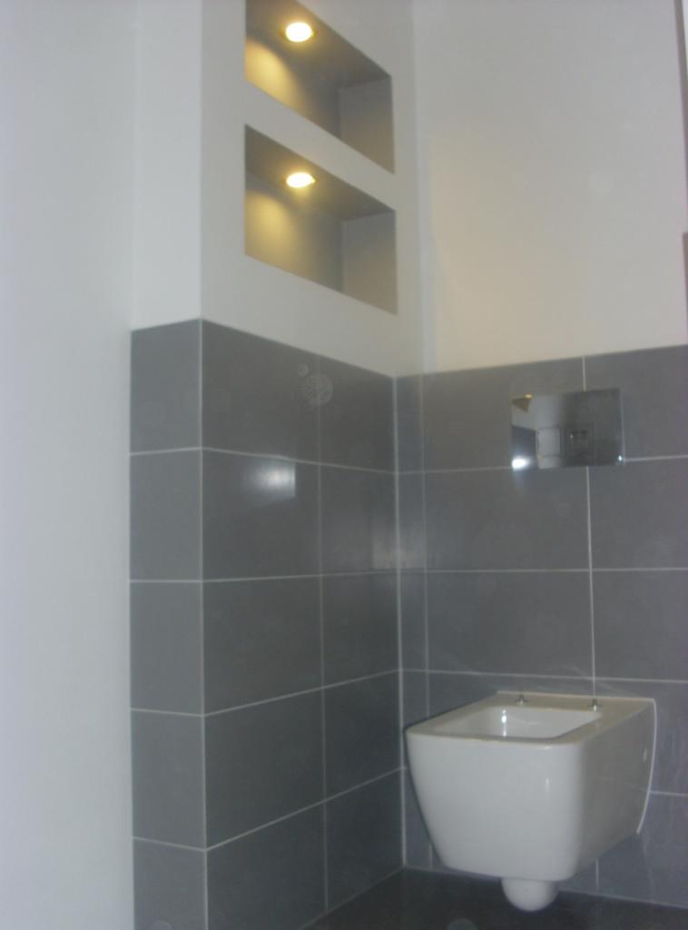 Szare płytki w łazience z podwieszanym bidetem i wnęką z oświetleniem punktowym. Minimalistyczny design, nowoczesne wykończenie.