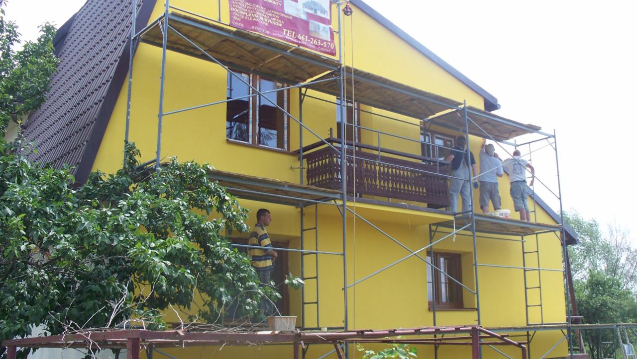 Żółty dom w trakcie malowania elewacji. Ekipa na rusztowaniu, widoczny fragment dachu i zieleń. Na elewacji baner reklamowy firmy.