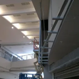 Montaż konstrukcji metalowej do płyty na ścianie wewnątrz budynku, widoczny podnośnik nożycowy i fragment sufitu z oświetleniem.