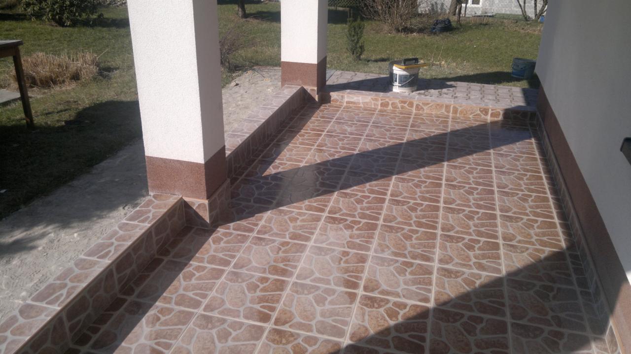 Zadaszony taras z brązowymi, wzorzystymi płytkami ceramicznymi, obramowany cokołem z tej samej płytki, z białymi filarami dekoracyjnymi z brązową podstawą, częściowo zacieniony.