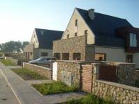 Dwa domy jednorodzinne z elewacją częściowo wykończoną kamieniem naturalnym, widoczne ogrodzenie z kamienia i drewnianych paneli, trawnik przy chodniku, w tle zaparkowany samochód osobowy.