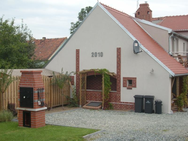 Jasna elewacja domu z czerwonymi ceglanymi akcentami, rok 2010 na ścianie, pergola obrośnięta bluszczem i murowany grill na tle ogrodzenia.