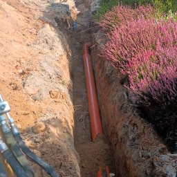 Usługi ogólnobudowlane - Pomarańczowe rury ułożone w wykopie ziemnym, widok z kabiny koparki, w tle pracujący robotnicy i budynek.