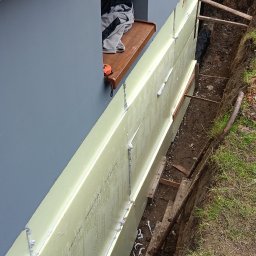 Usługi ogólnobudowlane - Fundament domu z izolacją termiczną, widoczne płyty styropianowe i wykop. Na parapecie leży miarka. Ściana w kolorze szarym, element dekoracyjny imitujący drewno.