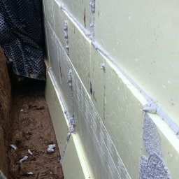Usługi ogólnobudowlane - Ocieplenie fundamentu z użyciem styropianu, zabezpieczone folią kubełkową. Widoczne łączenia płyt styropianowych wypełnione pianką montażową. Grunt wokół fundamentu.