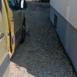 Żwirowa ścieżka obok budynku, widok z boku samochodu. Ścieżka prowadzi do bramy, w tle zabudowa jednorodzinna. Słoneczny dzień.