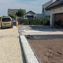 Wykończony podjazd z jasnego żwiru i białych kamyków oddzielonych szarym obrzeżem. W tle dom z garażem i zaparkowany samochód.