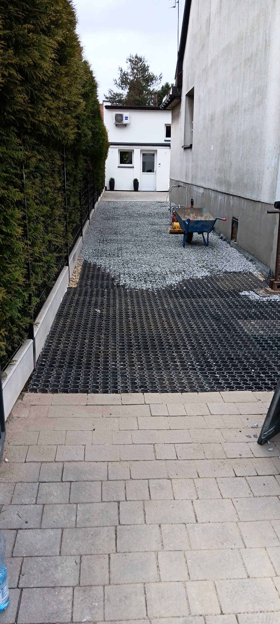 Ukończone utwardzenie terenu z użyciem plastikowych paneli i żwiru. Widoczna taczka i zagęszczarka, budynek w tle.