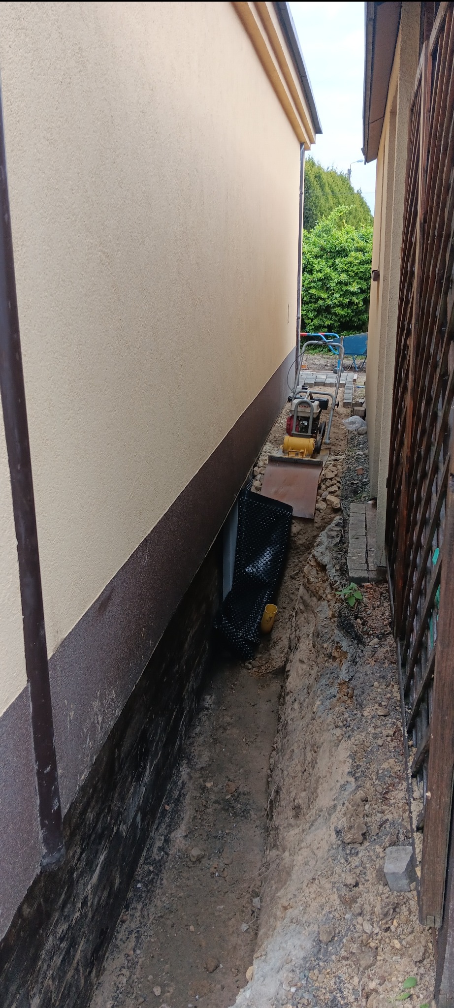 Wykopany wąski rów wzdłuż ściany budynku, częściowo zabezpieczony czarną folią kubełkową. W tle ubijarka wibracyjna i kostka brukowa. Prace ziemne.