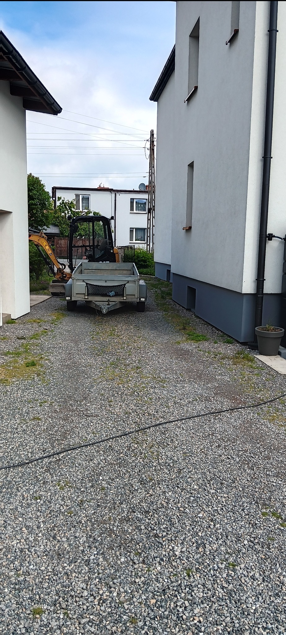 Mini koparka z przyczepką na żwirowym podłożu między dwoma budynkami. Widoczne instalacje elektryczne i fragmenty zieleni w tle.