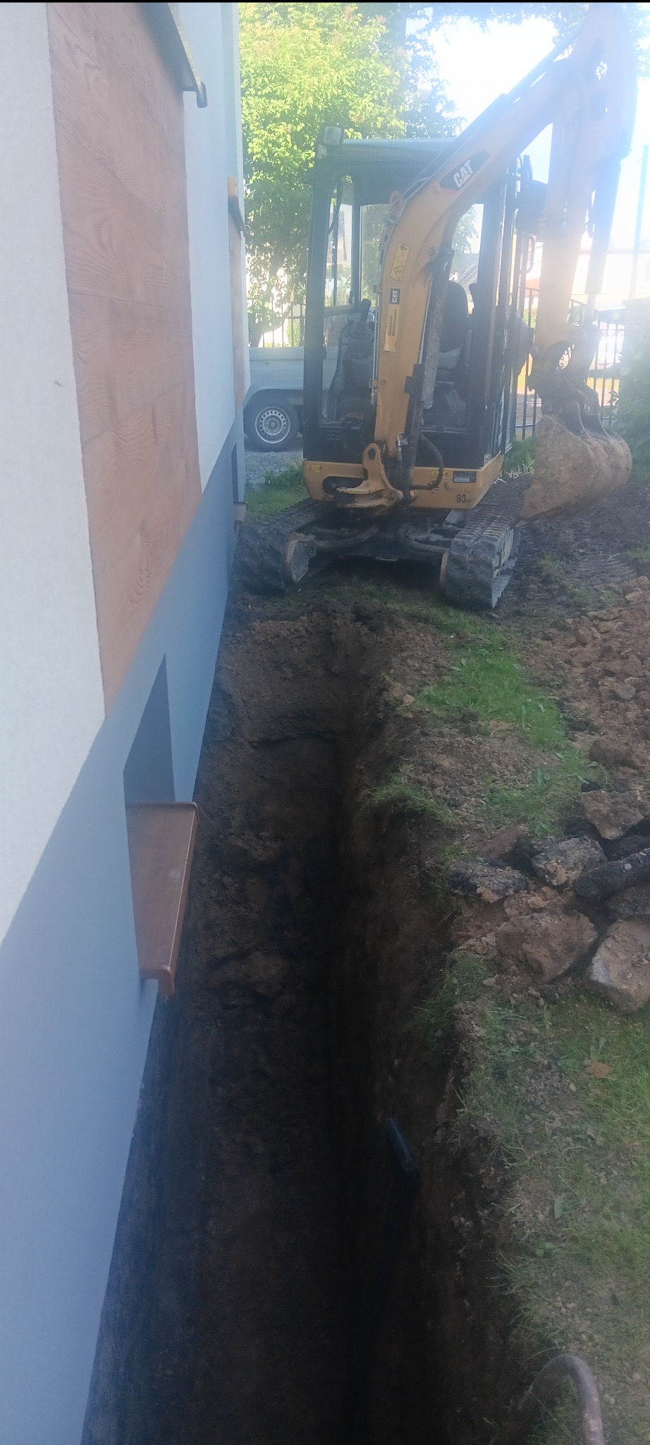 Wąski wykop wzdłuż ściany budynku, z widocznym małym koparką CAT. Wykop przygotowany pod izolację fundamentów. Widoczne ślady pracy koparki.