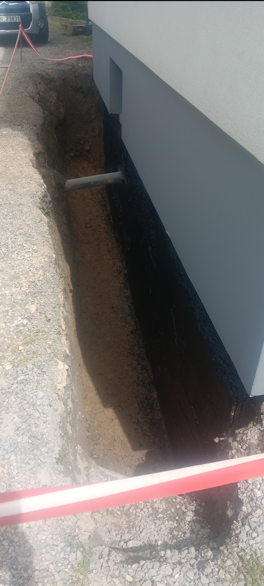 Fundament budynku po izolacji bitumicznej, widoczny wykop z rurą drenażową i taśmą ostrzegawczą.