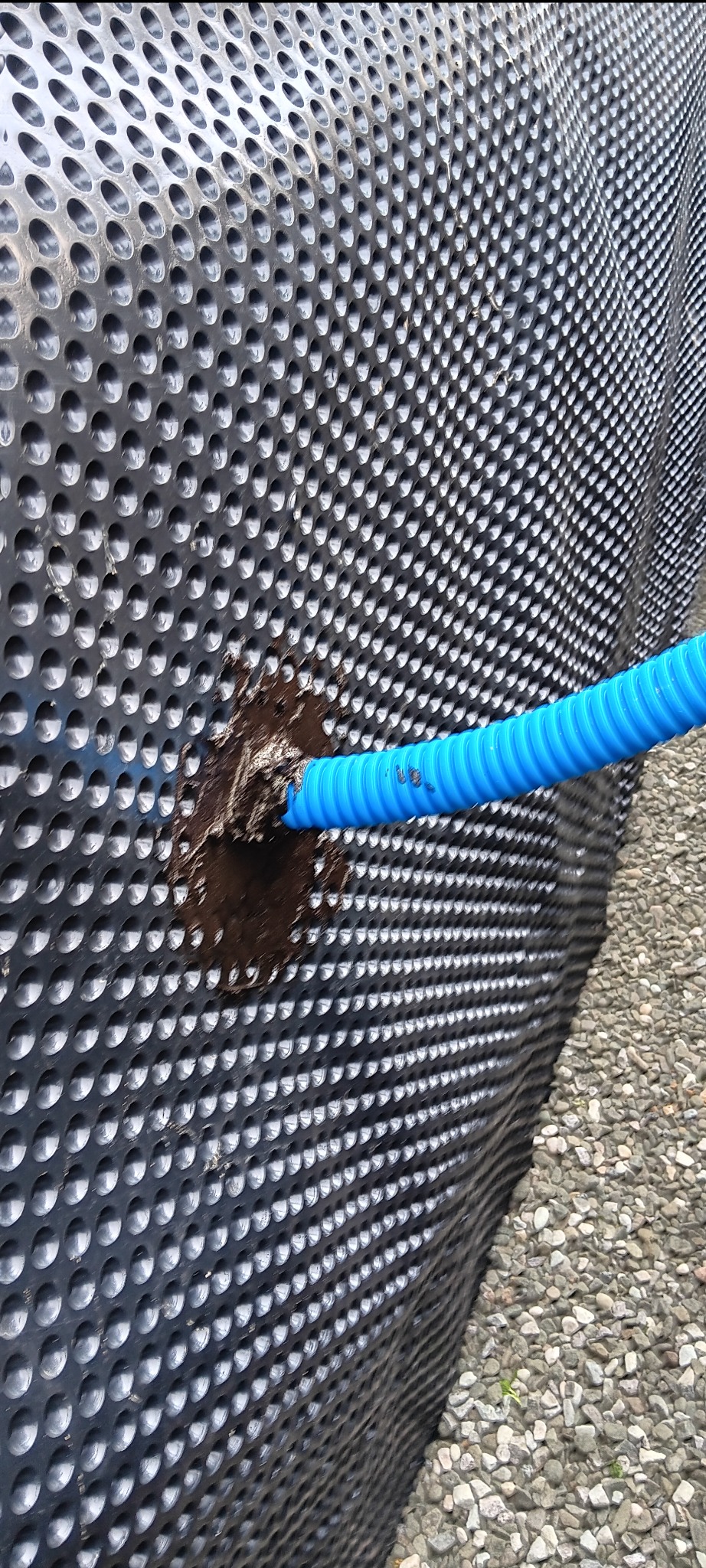 Czarna folia fundamentowa z wytłoczeniami i niebieska rura drenażowa wchodząca w otwór. Obok widoczny fragment podłoża z kamieni.