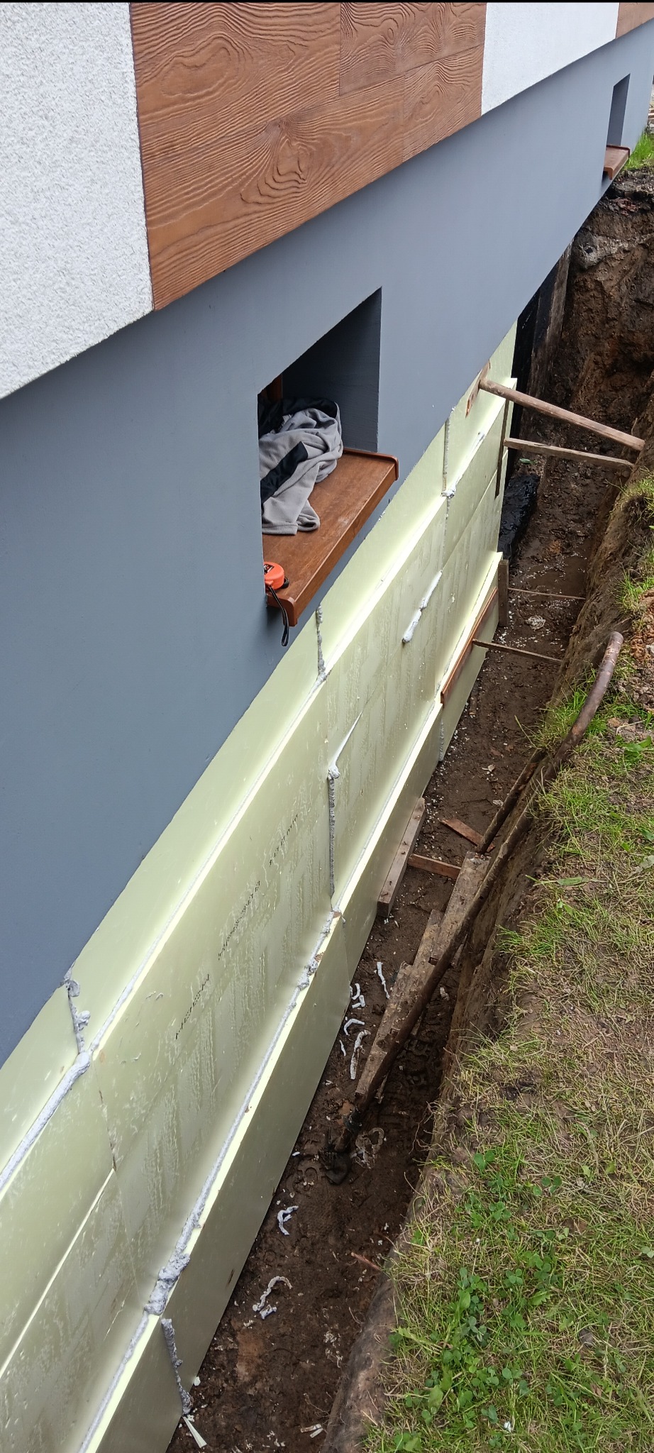 Fundament domu z izolacją termiczną, widoczne płyty styropianowe i wykop. Na parapecie leży miarka. Ściana w kolorze szarym, element dekoracyjny imitujący drewno.