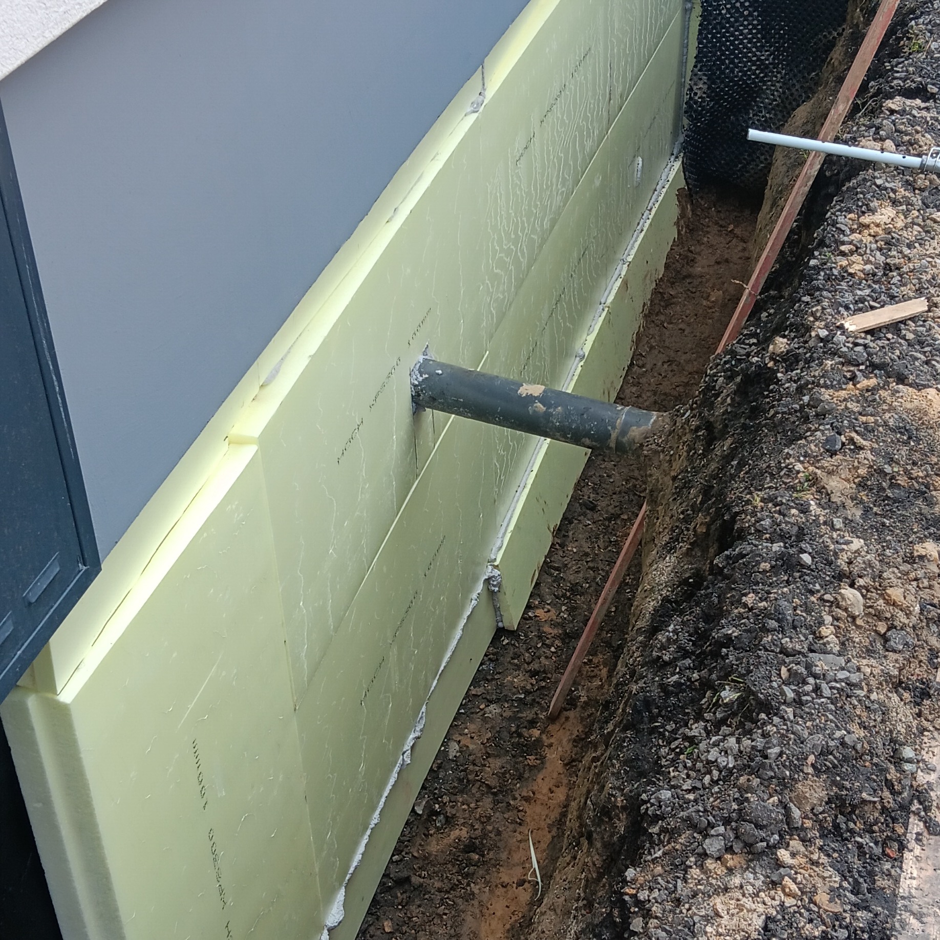 Żółta izolacja fundamentów z widoczną rurą drenażową, zabezpieczona czarną folią kubełkową, w wykopie z ziemią. Ujęcie z góry.